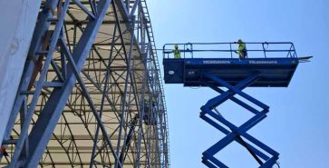 Ploščad z delavci ob zunanji strani industrijske hale med montažo konstrukcije.